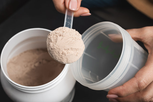 深色背景下，女性手将一勺巧克力乳清蛋白粉放入运动摇摇杯中，旁边是塑料蛋白粉罐，运动营养概念图片下载