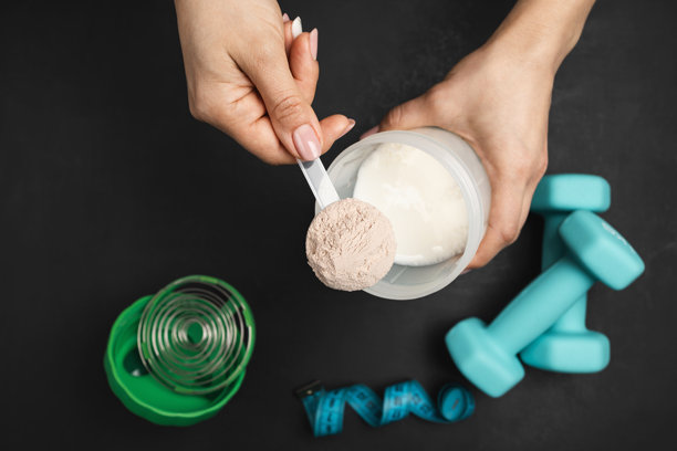 俯视角度拍摄的制作蛋白奶昔的画面，一只女性的手将一勺巧克力乳清蛋白粉放入装有牛奶的摇摇杯中，背景是深色的，并放有哑铃和卷尺图片下载