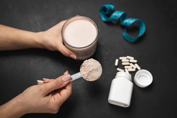 深色背景下，女性手持蛋白粉摇摇杯、一勺巧克力乳清蛋白粉、白色胶囊和卷尺，运动营养概念图片下载