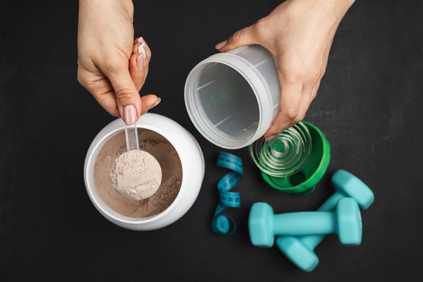 女性手从深色背景的蛋白粉罐、哑铃和卷尺旁，将一勺巧克力乳清蛋白粉放入摇摇杯中的俯视视角，运动营养概念图片下载