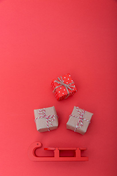 红色的雪橇和三个包装好的礼品盒放在红色背景上。圣诞节的喜悦、赠予和节日传统概念。竖版照片，俯视，平铺。图片下载