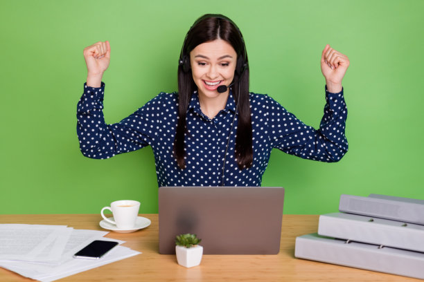 照片：一名年轻女子戴着耳机，拿着麦克风，穿着波点衬衫，在绿色背景前使用笔记本电脑，庆祝工作。图片下载