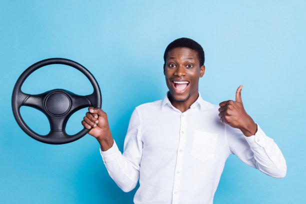 照片：一位穿着正装衬衫、肤色黝黑的帅气迷人男子，他竖起大拇指，微笑着驾驶汽车，背景是孤立的蓝色。图片下载
