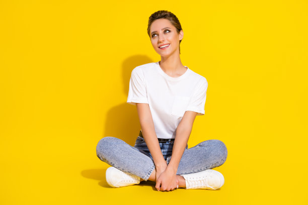 照片：可爱的、梦想般的女人，穿着休闲装，微笑着坐在地板上，望着空旷的空间，背景是孤立的黄色图片下载