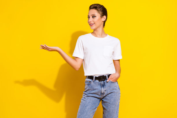 照片：乐观的棕色头发年轻女子站立，手持物品，望向空白空间，身穿白色T恤和牛仔裤，背景为黄色图片下载