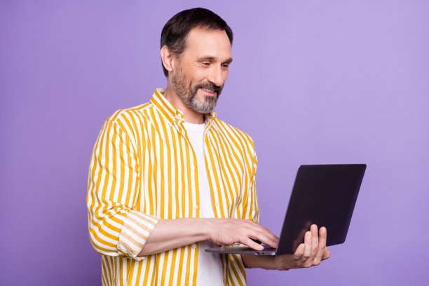照片肖像：穿着条纹衬衫的胡须男士，面带微笑，使用笔记本电脑工作，背景为孤立的淡紫色图片下载