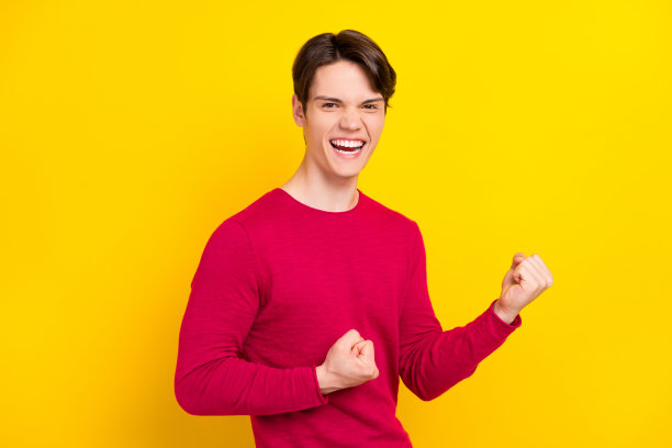 照片：欢呼的棕发千禧一代男性，双手握拳，穿着红色毛衣，孤立在黄色背景上图片下载