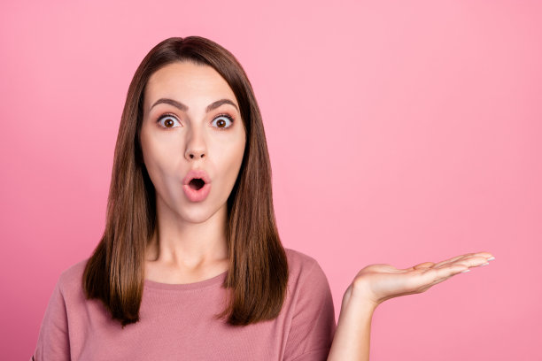 照片：印象深刻的棕色头发年轻女子穿着t恤，手持空白空间，孤立在粉红色背景上图片下载