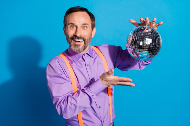 照片：一位快乐成熟的男士，带着积极的笑容，展示着手持迪斯科球，在蓝色的背景下庆祝派对图片下载