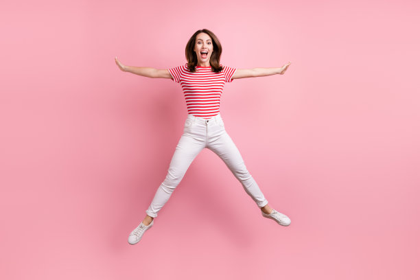 粉色背景上，年轻兴奋的女孩高高跃起，面带开心的笑容，如同明星一般，全长照片，孤立的。图片下载