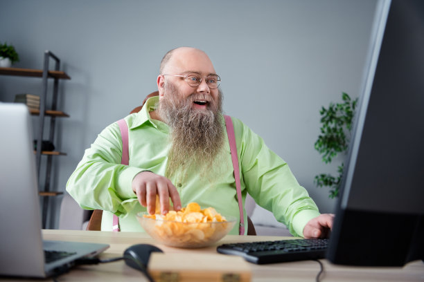 照片：一位快乐有趣的胖乎乎的胡子男坐在书桌前，在家庭办公室室内休息，吃着薯片零食看电影。图片下载