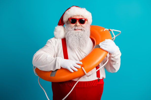 圣诞老人穿着红色吊带，手持救生圈，在蓝色背景前摆姿势，用于节日库存照片图片下载