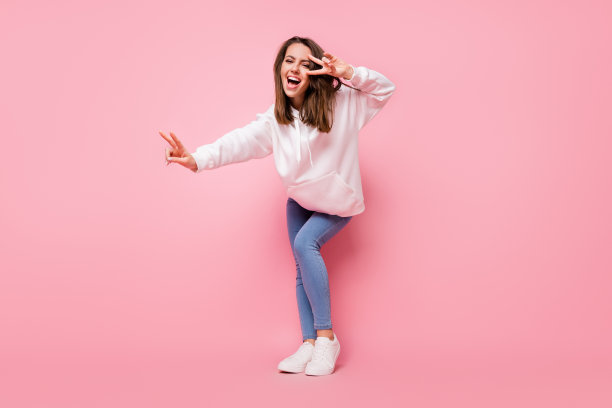 千禧一代的年轻女子，头发棕色，面带笑容，身穿白色连帽衫和牛仔裤，比划着V字手势，在粉色背景前跳舞，全尺寸照片图片下载