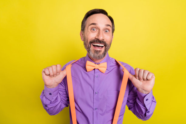 照片：一位穿着时髦、面带兴奋笑容的中年男性，拉着吊带，表情积极，在黄色背景前摆出有趣姿势图片下载