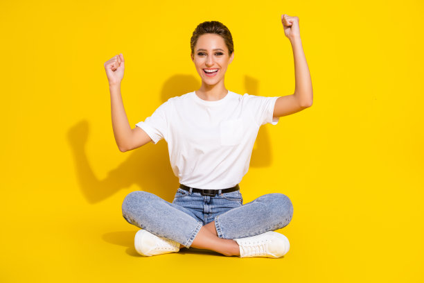 照片：一位幸运、时髦的年轻女士穿着休闲服，坐在地板上，握紧拳头，面带微笑，背景为孤立的黄色。图片下载