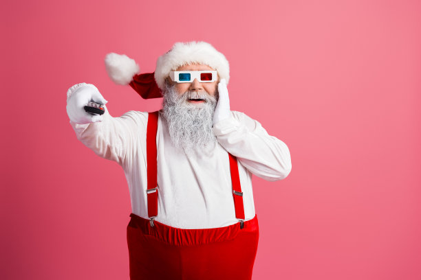 留着白色胡须、穿着红色吊带裤和3D眼镜的圣诞老人，手持遥控器，在粉色背景前俏皮地摆姿势，拍摄圣诞主题照片图片下载