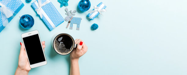俯视视角，女子一手持手机，一手持咖啡杯，背景为蓝色。圣诞装饰品和玩具。新年节日概念。样机图片下载