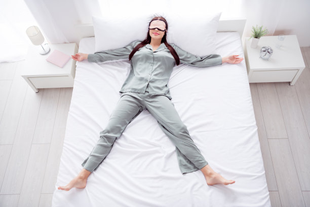 照片：一位穿着闪亮灰色睡衣的有趣年轻女子，戴着眼罩，面带微笑，在家里的房间里像星星一样睡着。图片下载