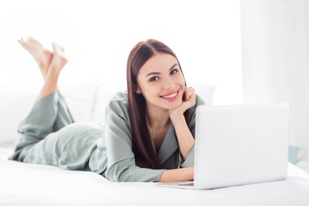 照片：一位穿着灰色睡衣的有趣、爱幻想的年轻女子，躺在床上，微笑着使用现代小工具，在室内房间、家中工作。图片下载