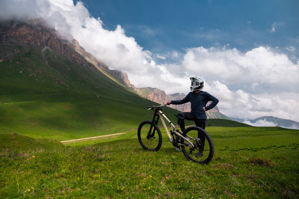 戴着头盔和眼镜的满意男子站在云层下的山地自行车旁，背景是覆盖着岩石山的云层天空。极限运动。图片下载