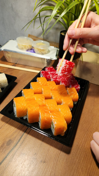 一只手用筷子从黑色托盘中夹起红色的寿司卷，托盘里还有橙色的三文鱼寿司，背景是木桌、绿植和其他寿司托盘，这是为日式餐饮菜单、外卖或餐厅拍摄的照片图片下载