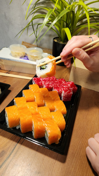手持筷子从黑色托盘中夹起一块红色寿司，托盘内有橙色寿司卷，背景有绿色植物，菜单特写照片，寿司外卖服务，日式餐厅，餐饮服务图片下载