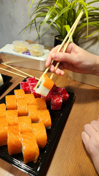 手持筷子从黑色托盘中夹起一块红色寿司，托盘里还有其他寿司卷，背景有绿色植物，菜单近景照片，寿司外卖服务，日式餐厅，餐饮服务图片下载