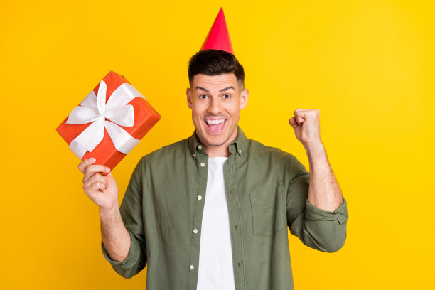 照片肖像，一个男人戴着生日头饰，微笑着拿着礼物盒，背景是鲜艳的黄色图片下载