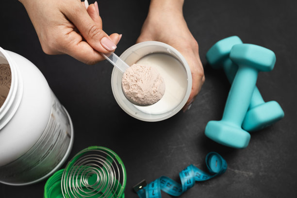 准备蛋白质奶昔，一只女性的手将一勺巧克力乳清蛋白粉放入装着牛奶的摇摇杯中，背景为深色，上面有蛋白质罐、哑铃和卷尺。图片下载