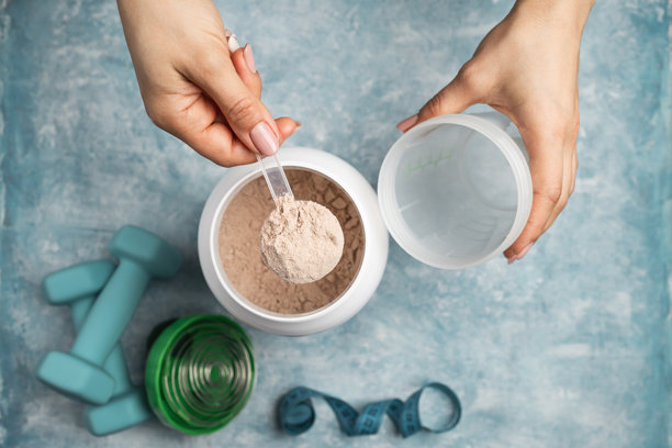 女性手部俯视视角，将一勺巧克力乳清蛋白粉放入运动摇摇杯中，背景为蓝色，配有蛋白粉罐、哑铃和卷尺，运动营养概念图片下载
