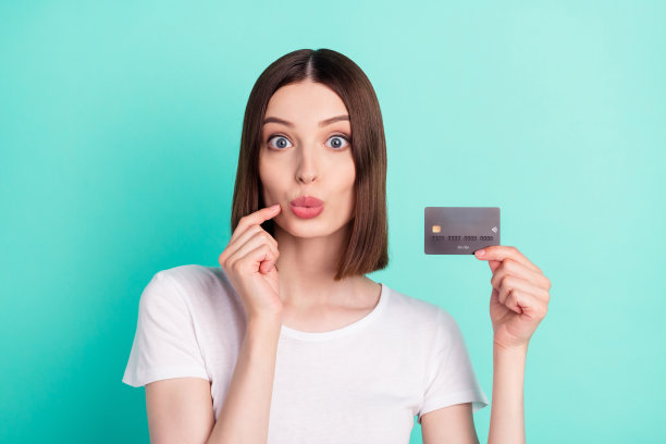 照片：年轻女子惊讶地捂住脸，手持信用卡支付，孤立在蓝绿色背景上图片下载