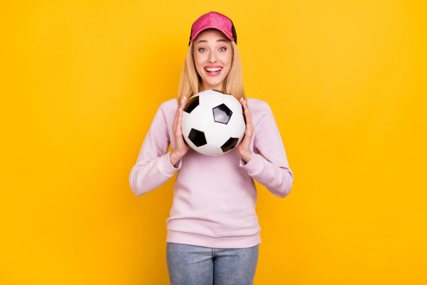 照片：戴着帽子、抱着足球、开怀大笑的快乐女人，在明亮的黄色背景前图片下载