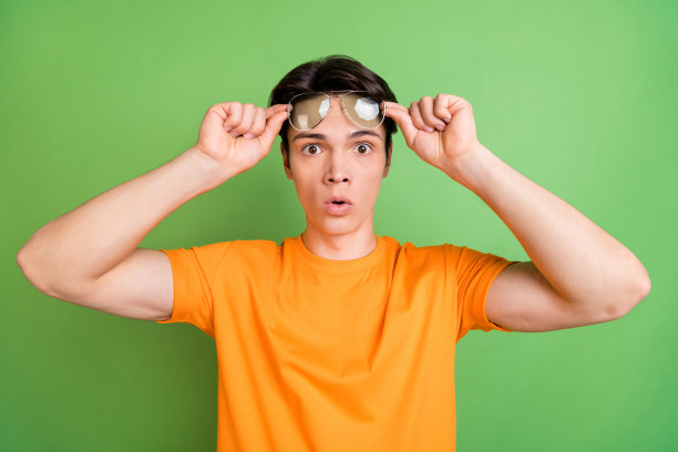 照片：戴着太阳镜、表情惊讶的年轻男子，双手捂脸，在绿色背景前做出新闻反应图片下载