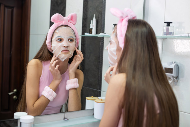 少女在浴室镜子前敷面膜。一位戴着发带和护腕的年轻女孩在护肤时，一边照镜子一边敷上保湿面膜。图片下载