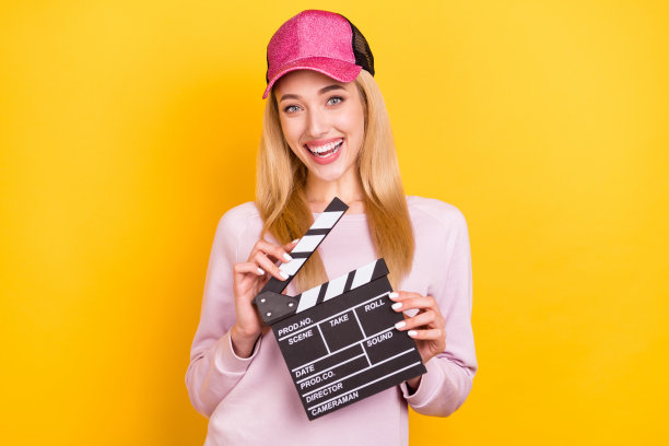 身着棒球帽的女子在明亮的黄色背景前，手持电影场记板，做出惊讶表情，进行电影拍摄，照片肖像图片下载
