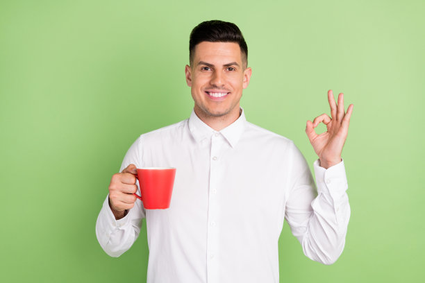 照片：英俊可爱的年轻男士穿着白衬衫，面带微笑，喝着饮料，竖起大拇指，背景为绿色。图片下载