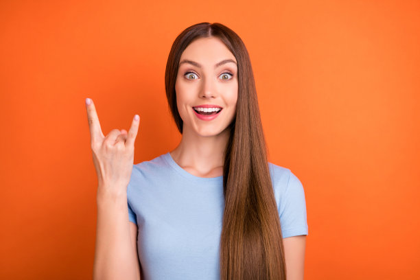 照片：一位留着长发、穿着蓝色T恤、展示摇滚手势的千禧一代女士，背景是鲜艳的橙色图片下载