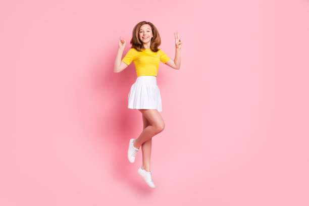 棕色头发的十几岁女孩身穿T恤和裙子，在粉色背景前跳跃并做出V字手势的全尺寸照片图片下载