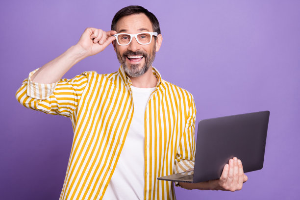 照片肖像：留着胡子的男人穿着条纹衬衫，戴着太阳镜，微笑着使用笔记本电脑，背景是孤立的淡紫色。图片下载