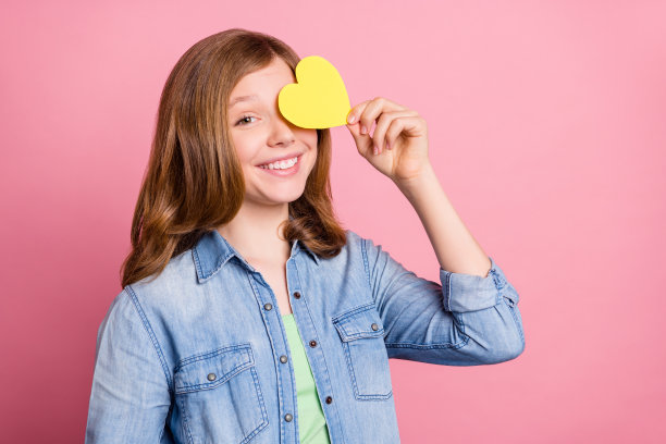 照片肖像：女孩微笑，用黄色心形明信片遮住眼睛，背景为孤立的柔和粉色。图片下载