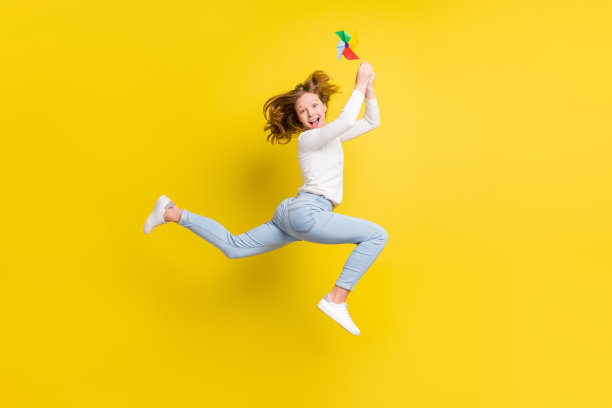 棕发小女孩穿牛仔裤，手持风车，在黄色背景下向上跳跃的全尺寸侧身照片图片下载