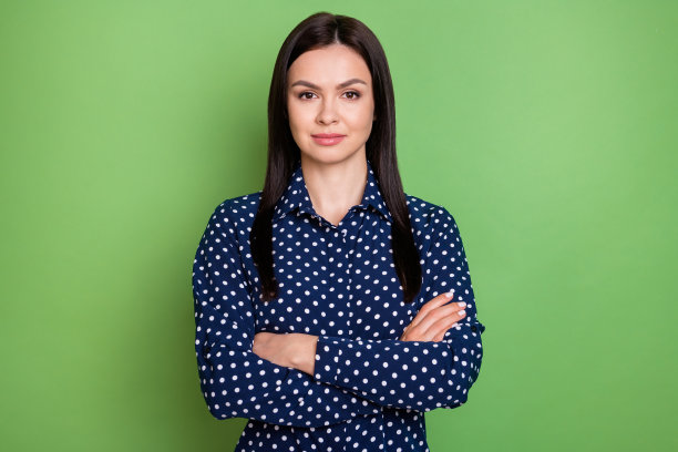 照片：一位留着棕色波浪卷发的强势千禧年女士，她双臂交叉，穿着蓝色衬衫，背景是绿色。图片下载