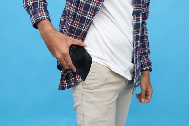 身穿休闲服的男子特写照片，他将手机放入裤子口袋，手机屏幕空白，背景为蓝色。图片下载