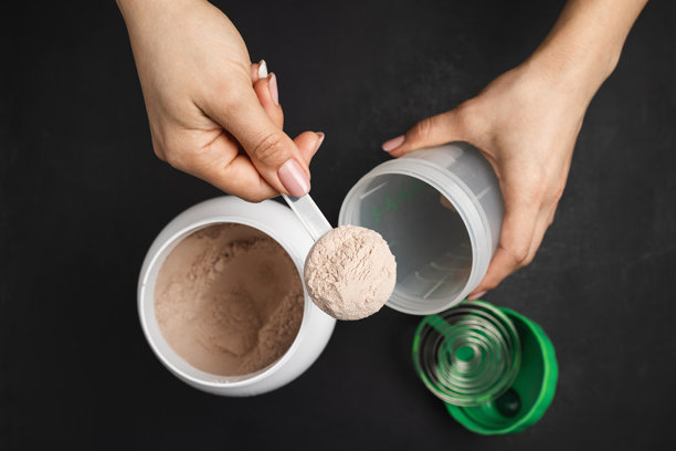 俯视女性手将一勺巧克力乳清蛋白粉放入运动营养概念的带塑料蛋白罐的摇摇杯中，背景为深色图片下载