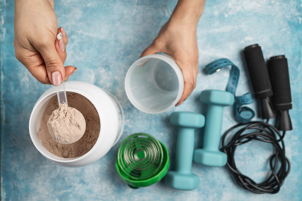 俯视视角，女性手将一勺巧克力乳清蛋白粉放入蓝色背景的摇摇杯中，旁边有罐子、哑铃、跳绳、卷尺，体现了健身准备和运动营养的概念图片下载