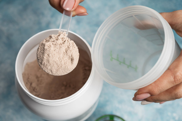 特写：女性手将一勺巧克力乳清蛋白粉放入蓝色背景的摇摇杯中，旁边是塑料蛋白粉罐，运动营养概念图片下载