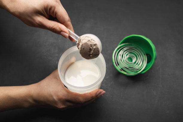 准备蛋白质奶昔，女性手将一勺巧克力乳清蛋白粉放入装有牛奶的摇摇杯中，背景为深色，运动营养概念图片下载