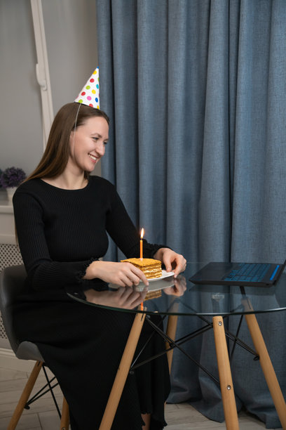 一位戴着派对帽的年轻女子在笔记本电脑前许愿，旁边放着生日蛋糕和蜡烛，正在线上庆祝生日。竖直照片图片下载