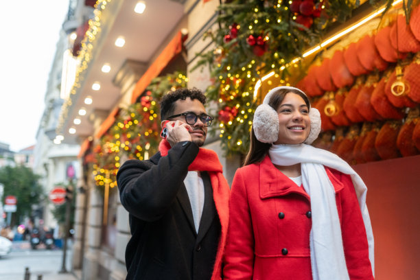 一对情侣在城市里庆祝节日季购物，男人在打电话，女人戴着耳罩。图片下载