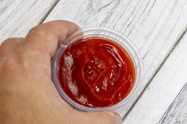 一只手拿着一杯透明的番茄酱，背景是纹理木板。这张图片捕捉到了调味品浓郁的红色，非常适合食品相关项目。图片下载